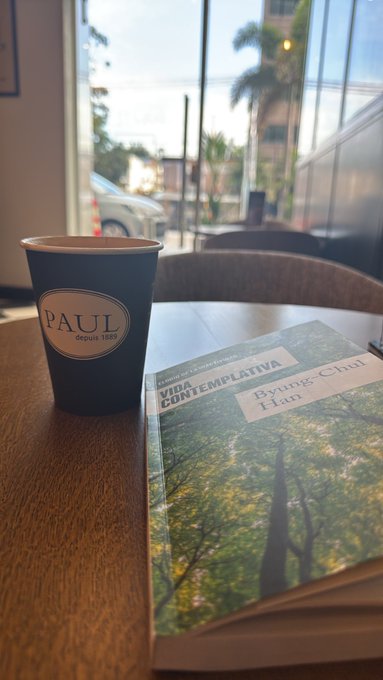 A coffee cup branded 'PAUL depuis 1889' sits on a table next to a book titled 'VIDA CONTEMPLATIVA' by Byung-Chul Han, photographed by a window.