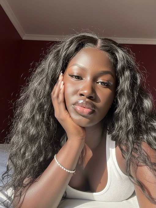 A close-up portrait shows a woman with long dark curly hair resting her hand on her cheek, wearing a white tank top and a bracelet, bathed in warm sunlight against a dark red wall.