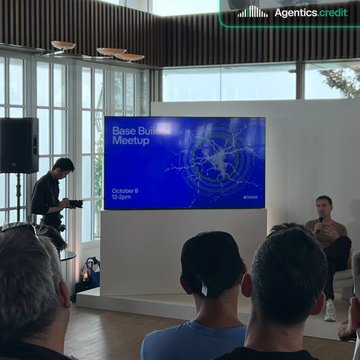 An audience watches a presentation at a 'Base Builders Meetup' displayed on a large screen in a modern venue with high ceilings and large windows.