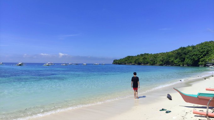 A person in red shorts walks along a sandy beach toward clear turquoise water with boats anchored nearby and a tree-covered coastline in the distance.