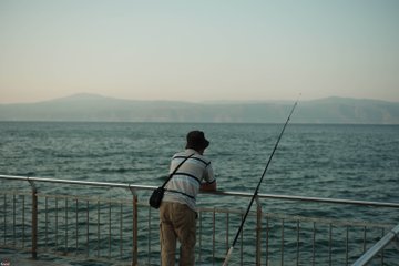 A person stands at a metal railing holding a fishing rod, looking out over the ocean towards distant mountains.