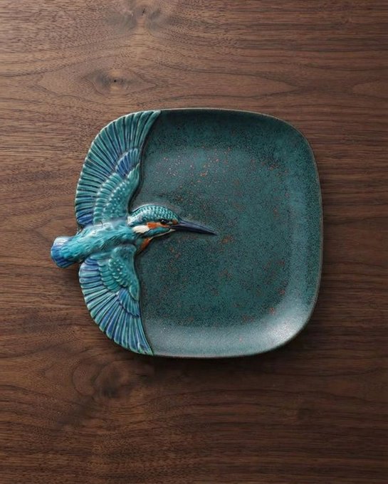 A decorative ceramic plate with a green speckled glaze and a sculpted kingfisher bird perched on the rim, resting on a wooden surface.