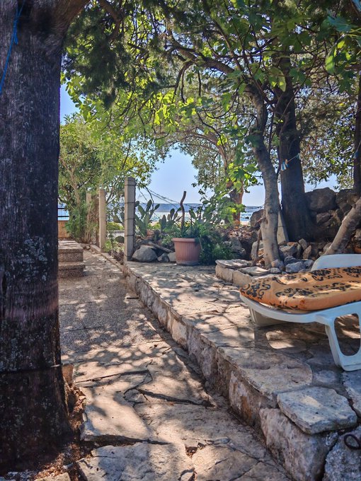 A peaceful garden pathway made of stone steps leading toward the sea, shaded by trees with a white lounge chair on the side.