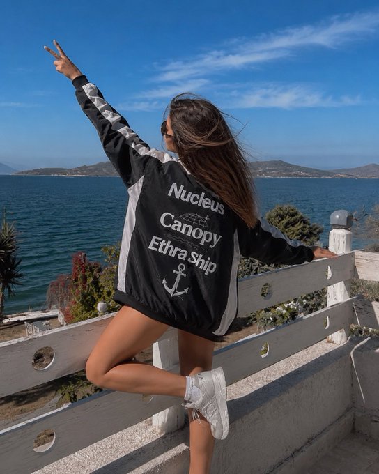 A woman standing on a white fence by the sea, wearing a black and white jacket with the text 'Nucleus Canopy Ethra Ship' and an anchor logo on the back, making a peace sign with her hand.