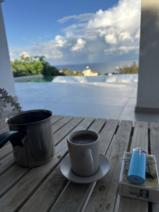 A coffee cup on a saucer, a stainless steel pitcher, a blue lighter, and a small box sit on a wooden slatted table overlooking an infinity pool and ocean horizon under a partly cloudy sky.