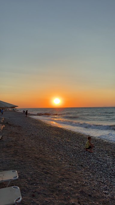 A serene sunset over the ocean at a pebbled beach, with a person sitting near the water and others walking in the distance.