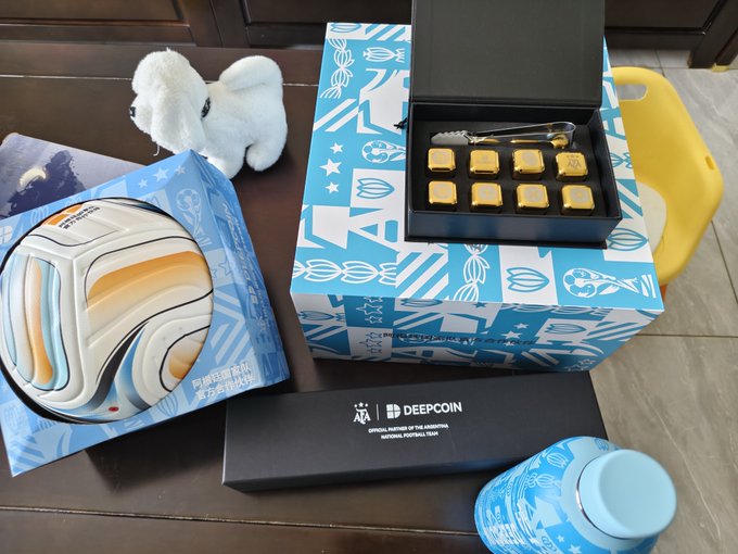 A promotional unboxing photo featuring FIFA World Cup 2026 merchandise, including a gift box with chocolates, a plush mascot, a 'DeepCoin' branded item, and a soccer ball design.