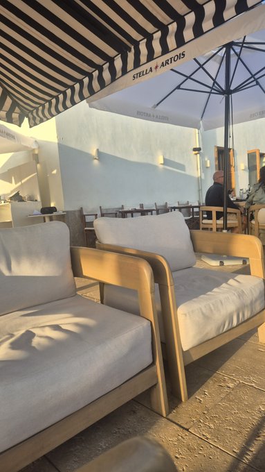 The image shows a patio area with two white chairs, one of which has an umbrella above it. The chairs are positioned under a striped awning that provides shade for the seating area. A person can be seen sitting at a table nearby, possibly enjoying their time in this outdoor space.