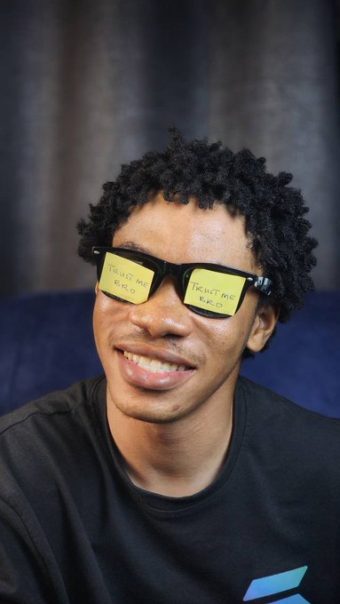 A young man smiles while wearing sunglasses with yellow 'TRUST ME BRO' stickers on the lenses.