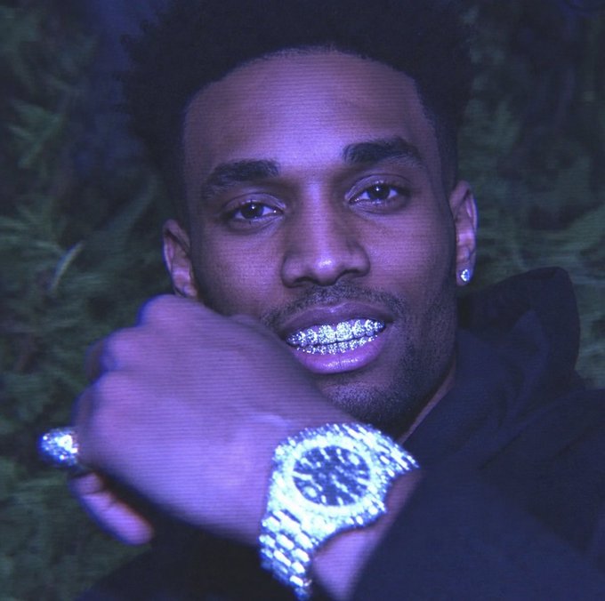 A close-up portrait of a young man with diamond-encrusted grillz on his teeth, holding his fist near his face to display a large, sparkling diamond-encrusted watch.