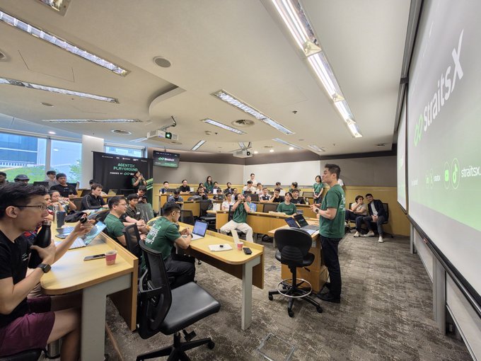 A large classroom or lecture hall is filled with attendees, many wearing green shirts, listening to a presenter standing near the front with a 'StratisX' banner displayed on a screen to the right.