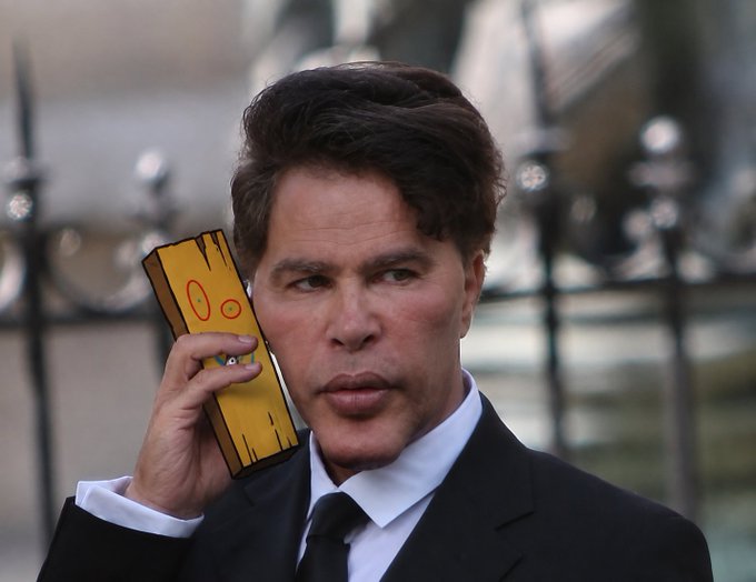 A man in a dark suit holds a yellow phone with a cartoon face on the case to his ear, featuring drawn-on eyes and a smiling mouth.