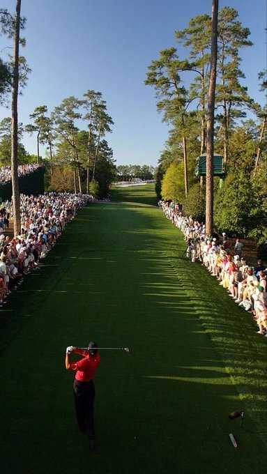 Tiger Woods, wearing a red shirt and black pants, follows through on a golf swing at the Masters Tournament while a massive crowd of spectators watches from behind the ropes.