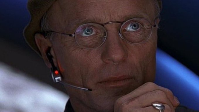 A close-up of an older man with glasses and a headset, displaying a shocked or stunned expression with wide eyes, likely a film still.