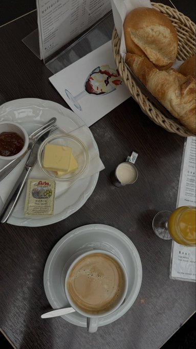 A coffee and croissant breakfast setup on a wooden table, featuring a basket of fresh bread, a cup of coffee, butter ("Bonnis"), jam, and a small pitcher of cream, perfectly paired for a relaxing morning to start the day.