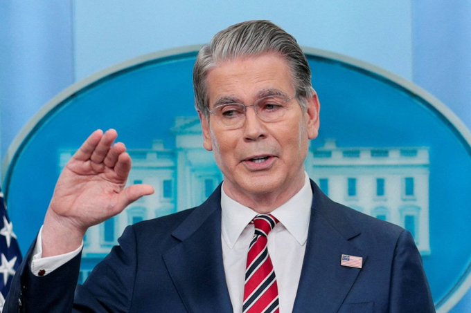 A man with glasses in a dark suit and red striped tie speaking at a podium with a White House backdrop, raising his hand in a gesture.