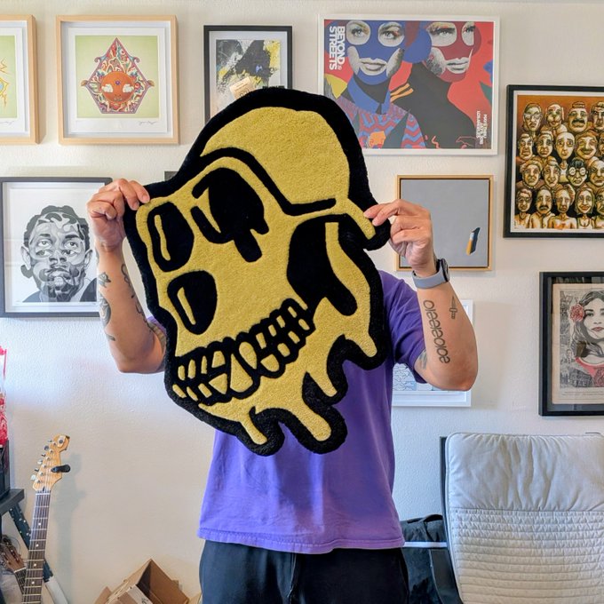 A person in a purple t-shirt holds up a large gold and black skull-shaped rug, obscuring their face, with framed art and a guitar visible in the background.