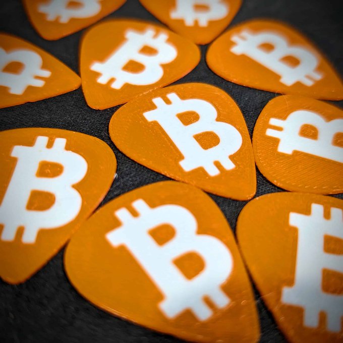 Multiple orange guitar picks featuring the white Bitcoin "₿" logo are arranged on a dark surface, visually representing the 10-pack of Bitcoin-themed guitar picks referenced in the post.