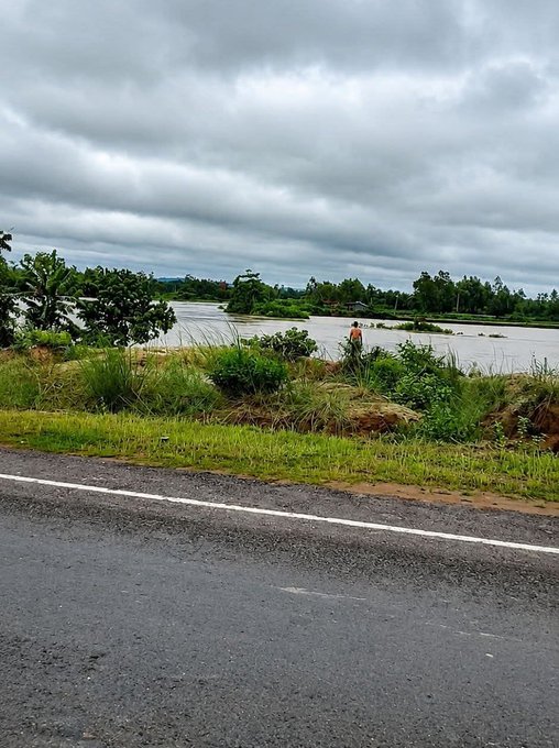 A moody landscape photo of a person standing by a river under an overcast sky, viewed from across a wet asphalt road with grassy banks.