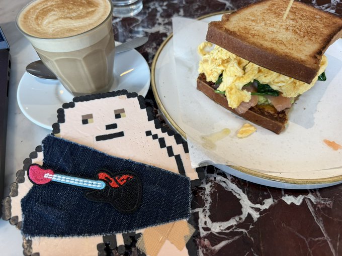 A latte and an egg sandwich on toast are served at a marble table alongside a pixel-art beaded bag depicting a character with a pink guitar.