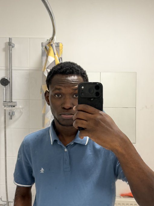 A man in a blue polo shirt takes a mirror selfie in a bathroom using a black phone.