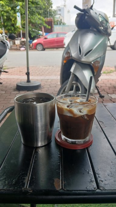 Two iced coffee drinks, one in a stainless steel cup and one in a glass with a straw, sit on a black outdoor table with a silver scooter and street visible in the background.