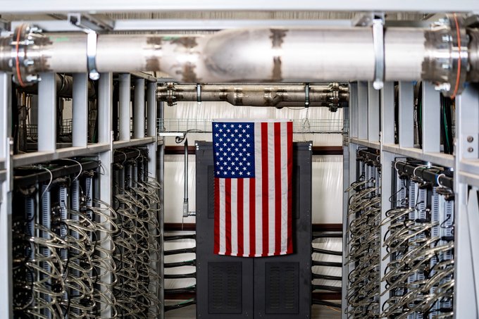An American flag hangs in the corridor between two rows of networking equipment racks filled with cables in what appears to be a data center.