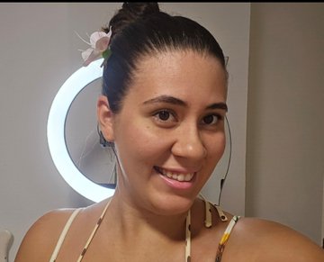 A smiling woman with dark hair tied up and a flower accessory looks at the camera in a selfie, with a ring light visible in the background.