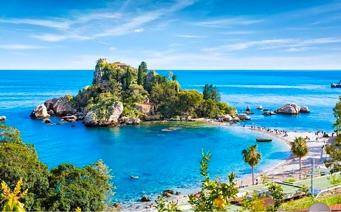 A scenic view of a rocky island topped with lush greenery and buildings, surrounded by bright turquoise water and a sandy beach dotted with palm trees.