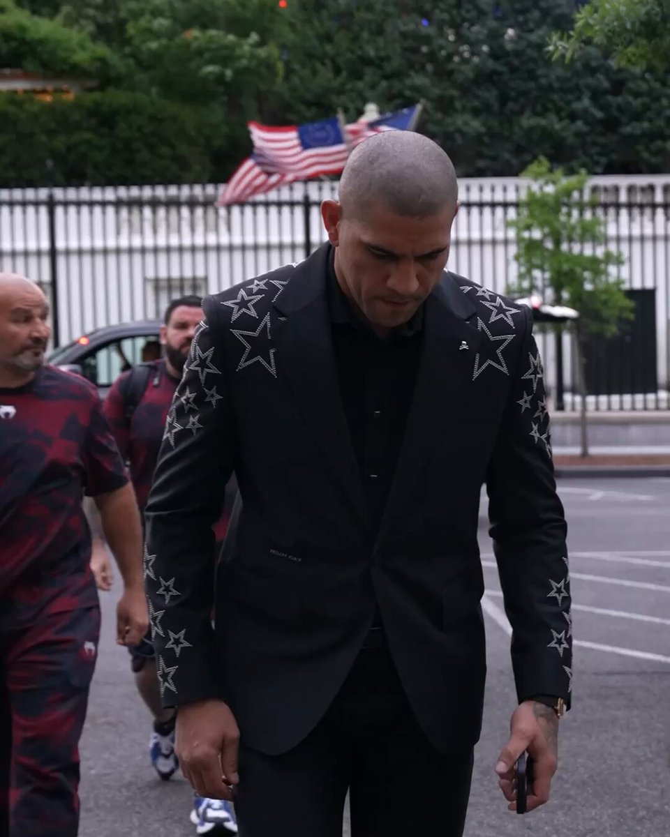 The image features a man wearing a black suit with white stars, walking down a street while holding a cell phone. The surrounding area includes trees and cars, indicating an urban setting. The caption of the image reads "For GOLD UFC Freedom [Title fights presented by @Cryptocom]". This suggests that the photo is related to a post about UFC fights or UFC fighter coverage on social media platforms.

The man in the suit appears to be walking alone, possibly heading towards a specific event or location. The presence of trees and cars in the background adds an interesting contrast between nature and urban life.