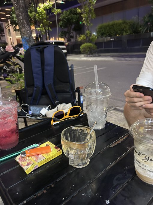 A nighttime outdoor cafe scene showing a black table with a backpack, sunglasses, an orange lighter, cigarette packs, and several drinks including a red beverage, an iced coffee, and a clear cup with a straw.