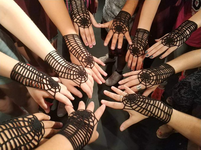 A group of people stand in a circle holding out their hands, all wearing matching black crocheted fingerless gloves and arm warmers with a spiderweb pattern.