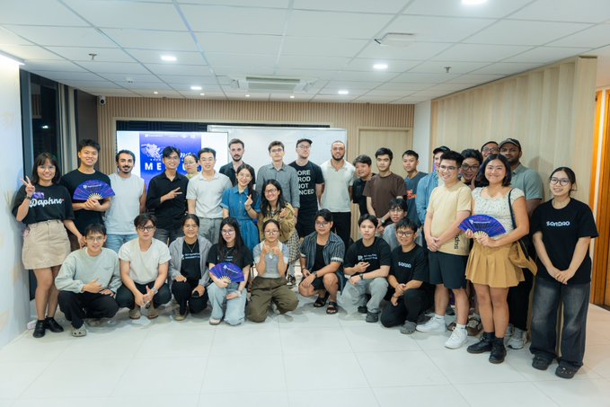 A large group of people poses for a group photo in an office hallway, with several individuals holding purple MoonDAO fans and wearing matching t-shirts.