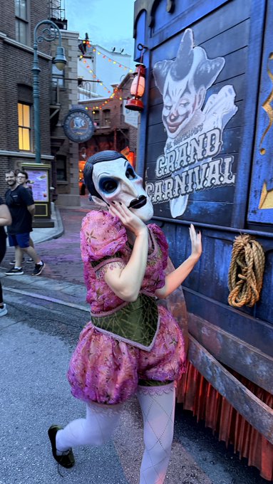 A person in a pink dress and theatrical mask poses next to a wooden structure with a 'Graine's Graine Carnival' painted sign, set in a themed outdoor street environment.