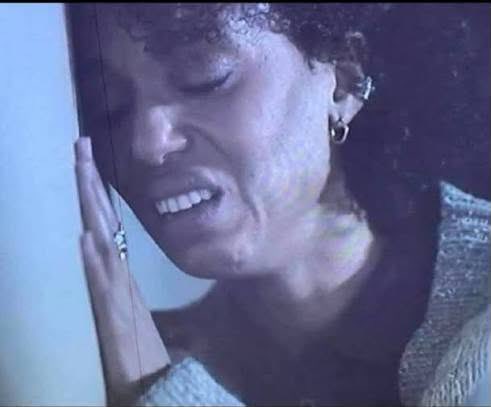A vintage-style, moody close-up photo of a woman smiling with her eyes closed and hand pressed against a wall, appearing emotional or relieved.
