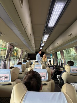 Interior view from the back of a tour bus showing passengers seated in beige chairs with personal entertainment screens, with a standing guide or speaking person visible near the front of the cabin.