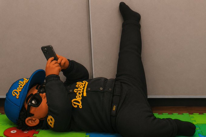 A child wearing a blue "Decibel" cap and black jacket lies on a colorful play mat, looking at a smartphone while leaning against a wall.