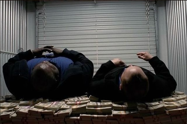 Two men in dark suits lie on top of large stacks of banded US currency inside a vault-like room with metal blinds.