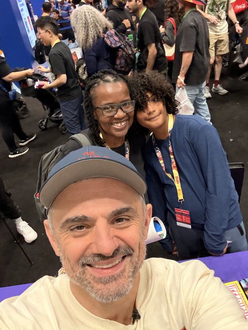 A selfie taken by a smiling bald man wearing a "THE TONIGHT SHOW" baseball cap, posing with a smiling woman in glasses and a curly-haired boy in a blue sweatshirt, in what appears to be a convention setting.