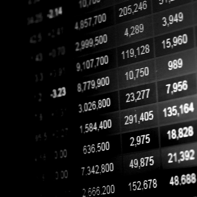 A close-up, angled view of a black digital stock market ticker board displaying rows of numerical financial data in white text.