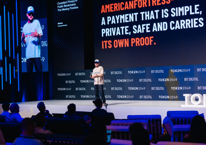 A man gives a presentation on stage at a Token2049/Bit Digital conference, with the slide showing 'AMERICANFORTRESS' and the text 'A PAYMENT THAT IS SIMPLE, PRIVATE, SAFE AND CARRIES ITS OWN PROOF.'