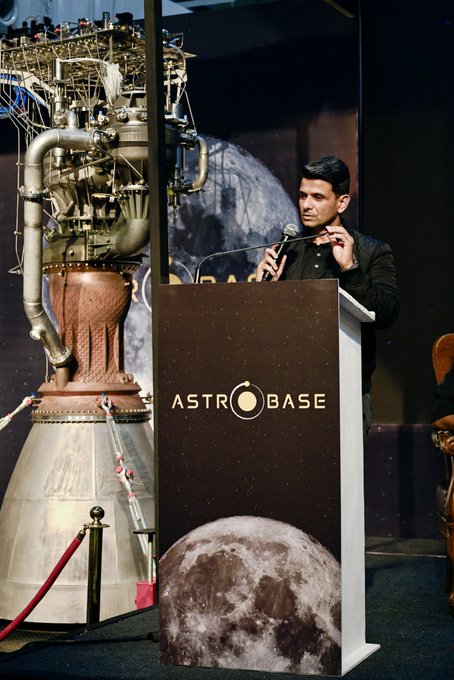 A man speaks at a podium branded 'ASTROBASE' while a large rocket engine model is displayed behind him, with a moon graphic on the podium front.