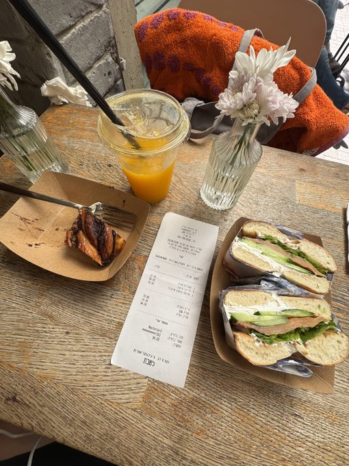 A wooden table displays a receipt, orange juice, a pastry, a sandwich cut in half, and a vase with pink and white flowers.