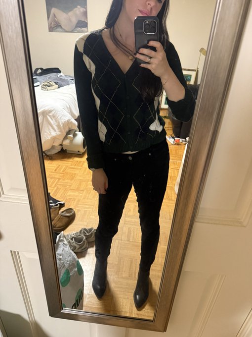 A person takes a mirror selfie wearing a black and white argyle cardigan, dark jeans, and black boots, standing in a bedroom with a bed and scattered shoes visible in the reflection.