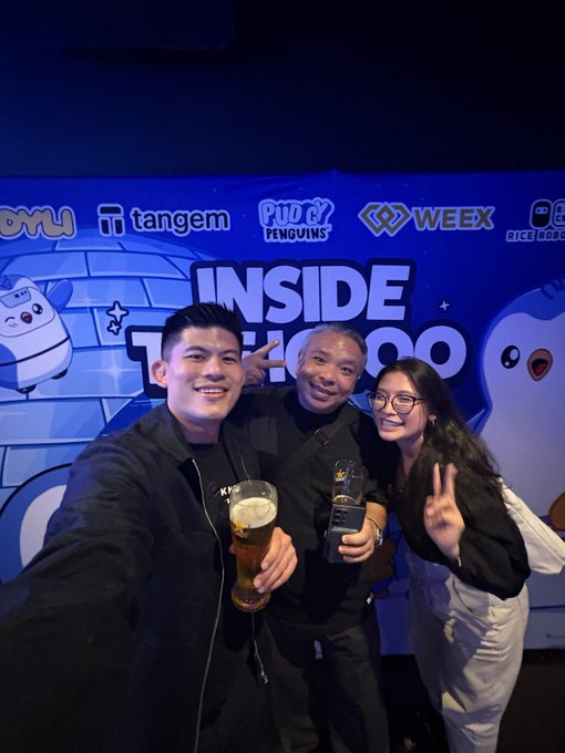 Three people pose for a selfie smiling and holding drinks in front of a blue backdrop featuring logos like Tangem, Pudgy Penguins, and WEEX with the text 'INSIDE' partially visible.