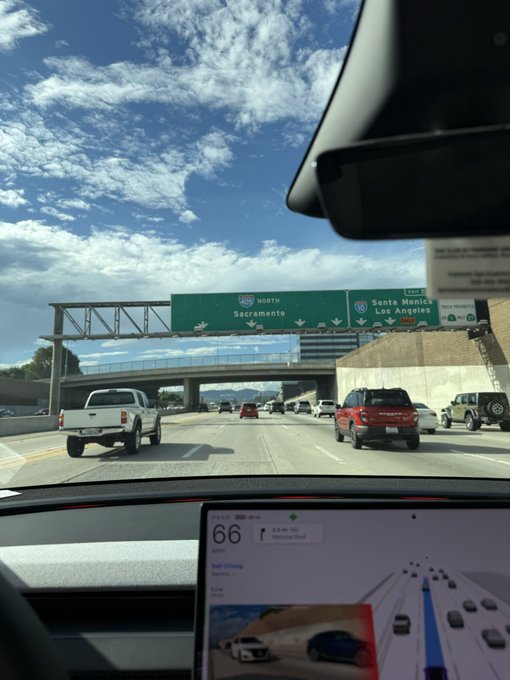 A view from inside a Tesla on a highway shows the dashboard screen displaying a speed of 66 mph, with overhead green road signs indicating directions for Sacramento (North 99) and Santa Monica/Los Angeles (East 10).