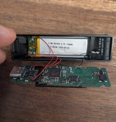 A person holds an opened Ledger hardware wallet device, exposing its internal circuit board and lithium polymer battery (PK501434, 3.7V, 700mAh) with a USB connector visible at the bottom, illustrating the teardown of a potentially compromised cryptocurrency security device.