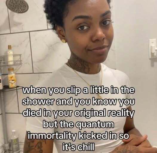 A young woman with tattoos takes a selfie in a bathroom with marble tile, wearing a white t-shirt and gold stud earring. Overlaid text reads: "when you slip a little in the shower and you know you died in your original reality but the quantum immortality kicked in so it's chill."