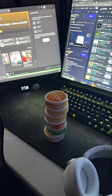 A stack of four Tropicana orange juice containers sits on a black desk next to a keyboard and dual monitors displaying Spotify and a trading platform, with white headphones in the foreground.