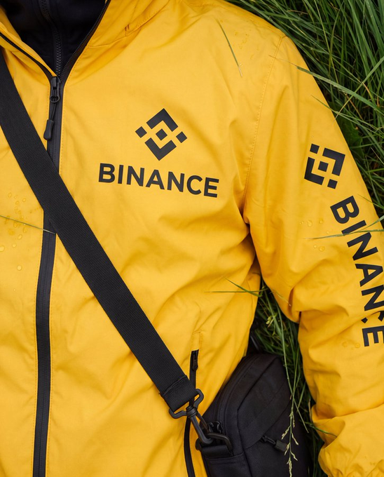 A bright yellow jacket featuring the "BINANCE" logo and branding lies in tall green grass, accompanied by a black crossbody bag—fitting the caption's "roll-on-grass kinda day" vibe with its laid-back outdoor aesthetic.
